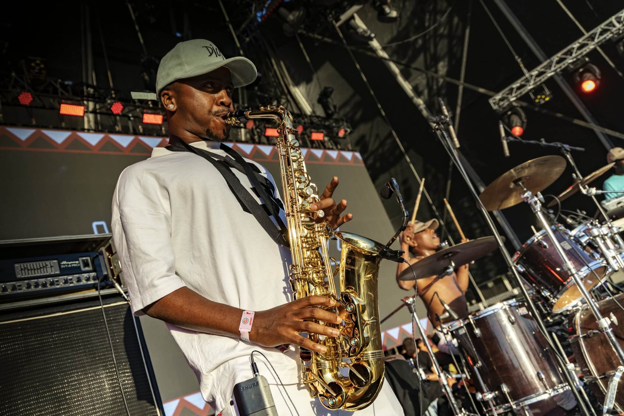 A Black male musician wearing a light-colored t-shirt and a baseball cap playing a tenor saxophone on a stage during a performance at African music festivals